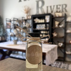 May include: A clear glass bottle with a black cap and a brown label, tied with twine. The label reads "Rush River Candles" and features a botanical design. The background includes the text "Rush River" on the wall.