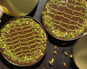 May include: Two round pistachio kunafa dream cakes topped with chocolate and pistachio sprinkles. The cakes are in gold-colored containers on a wooden surface. The text "Pistachio Kunafa Dream Cake" is written below the cakes.