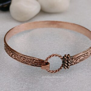 May include: A copper-colored bangle bracelet with an intricate floral pattern along the edges. The bracelet features a twisted rope clasp and a decorative floral design. The bracelet is approximately 2.5 inches in diameter.