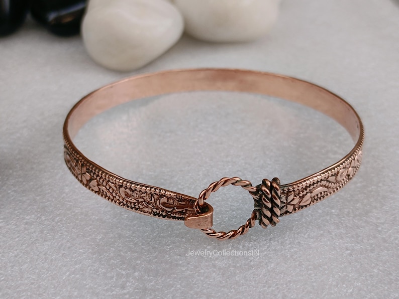 May include: A copper-colored bangle bracelet with an intricate floral pattern along the edges. The bracelet features a twisted rope clasp and a decorative floral design. The bracelet is approximately 2.5 inches in diameter.