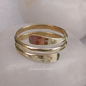 May include: A gold-coloured spiral ring with a hammered texture. The ring wraps around the finger multiple times, creating a layered effect. The ring is displayed on a white, rounded surface.