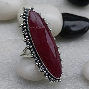 May include: A silver ring featuring a large, oval-shaped, dark red gemstone set in a silver bezel. The ring has a decorative border of small silver beads.