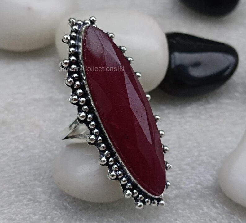 May include: A silver ring featuring a large, oval-shaped, dark red gemstone set in a silver bezel. The ring has a decorative border of small silver beads.