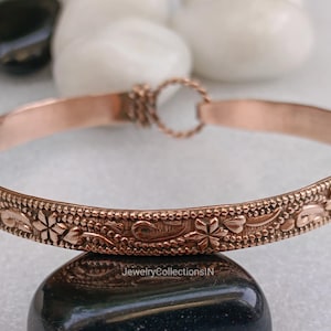 May include: A copper-colored bangle bracelet with an intricate floral design. The bracelet features a repeating pattern of flowers and leaves along its outer edge. The clasp is a small, decorative ring. The bracelet is displayed on a dark, rounded stone.