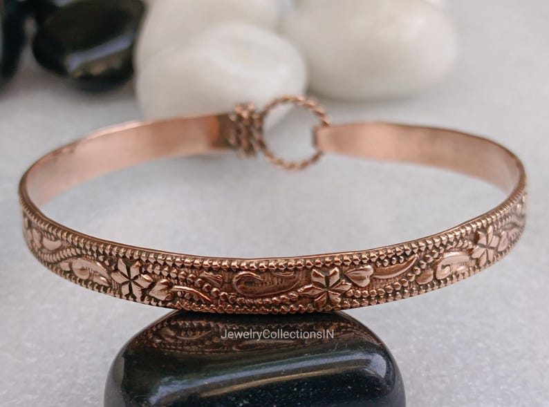 May include: A copper-colored bangle bracelet with an intricate floral design. The bracelet features a repeating pattern of flowers and leaves along its outer edge. The clasp is a small, decorative ring. The bracelet is displayed on a dark, rounded stone.