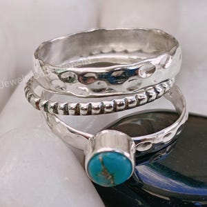 May include: A stack of silver rings. The top ring has a hammered texture, the middle ring has a beaded design, and the bottom ring features a turquoise stone. The rings are displayed on a white and black surface.