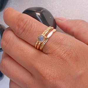 Puede incluir: Un conjunto de tres anillos dorados. Un anillo presenta una pequeña gema redonda de color azul claro. Los otros dos anillos tienen bandas texturizadas y lisas. Los anillos están apilados en un dedo.