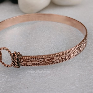 May include: A copper-colored bangle bracelet with an intricate floral and dotted pattern. The bracelet has a clasp closure and is approximately 2.5 inches in diameter. The jewelry piece is displayed on a light gray surface.