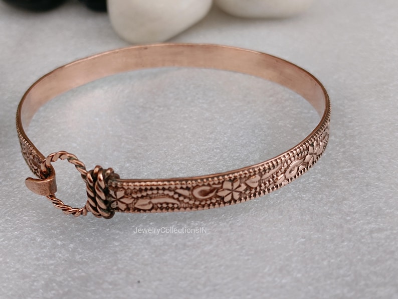 May include: A copper-colored bangle bracelet with an intricate floral and dotted pattern. The bracelet has a clasp closure and is approximately 2.5 inches in diameter. The jewelry piece is displayed on a light gray surface.