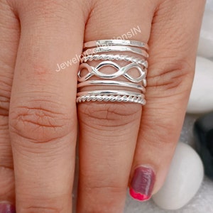 Puede incluir: Un conjunto de cinco anillos de plata apilados en un dedo. Los anillos son de diferentes estilos, incluyendo una banda martillada, un diseño de cuerda y un símbolo de infinito.