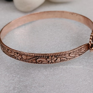May include: A copper-colored bangle bracelet with a floral design. The bracelet has an intricate pattern of flowers and leaves along the band. The clasp is a twisted rope design. The bracelet is approximately 2.5 inches in diameter.