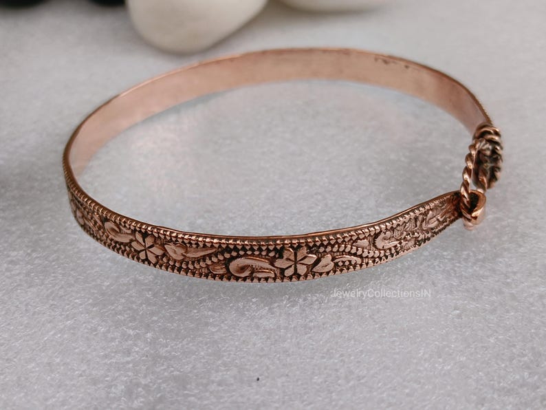 May include: A copper-colored bangle bracelet with a floral design. The bracelet has an intricate pattern of flowers and leaves along the band. The clasp is a twisted rope design. The bracelet is approximately 2.5 inches in diameter.
