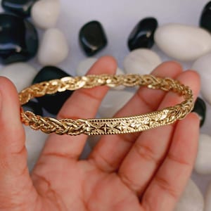 May include: A gold-colored bracelet with a braided design and a decorative central band featuring star-like patterns. The bracelet is held in a hand, showcasing its intricate details and craftsmanship. The background includes black and white stones.