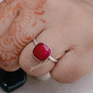 May include: A silver ring with a square-shaped, deep red gemstone. The ring is on a finger with henna.