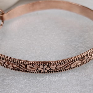 May include: A copper-colored bangle bracelet with an intricate floral pattern. The bracelet features a detailed design of flowers and leaves, with a textured border. The clasp is a simple loop and hook closure. The bracelet is approximately 2.5 inches in diameter.