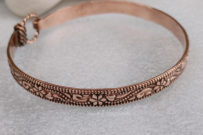 May include: A copper-colored bangle bracelet with an intricate floral pattern. The bracelet features a detailed design of flowers and leaves, with a textured border. The clasp is a simple loop and hook closure. The bracelet is approximately 2.5 inches in diameter.
