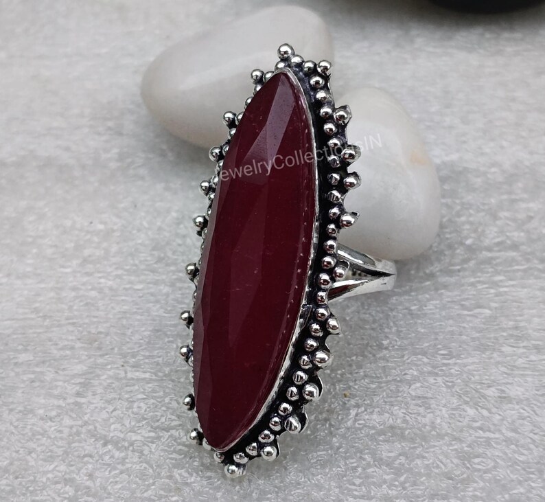 May include: A silver ring featuring a large, oval-shaped, dark red gemstone set within a decorative, beaded border.