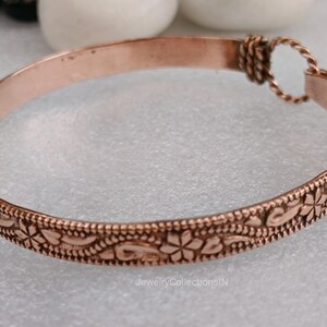 May include: A copper-colored bracelet with an engraved floral pattern. The bracelet has a simple, elegant design with a textured surface. The clasp is a twisted loop. The bracelet is approximately 0.5 inches wide.