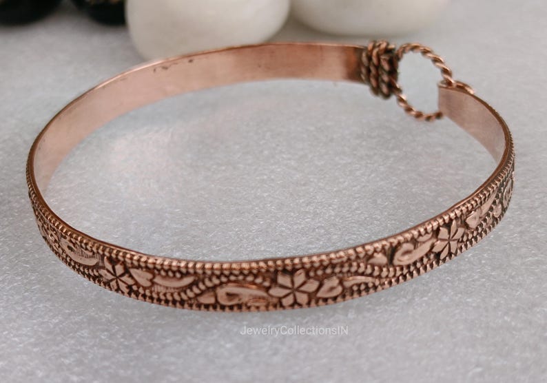 May include: A copper-colored bracelet with an engraved floral pattern. The bracelet has a simple, elegant design with a textured surface. The clasp is a twisted loop. The bracelet is approximately 0.5 inches wide.