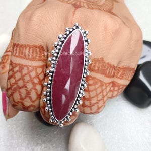 May include: A silver ring featuring a large, oval-shaped, dark pink gemstone. The ring has a decorative, beaded border.