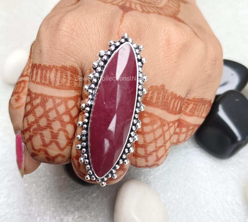 May include: A silver ring featuring a large, oval-shaped, dark pink gemstone. The ring has a decorative, beaded border.