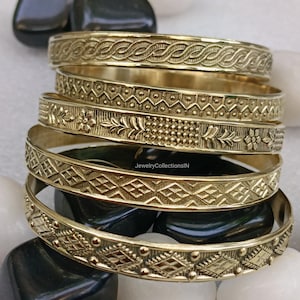 May include: A stack of six gold-colored bangles with intricate embossed designs. The bangles feature patterns like woven textures, geometric shapes, and floral motifs. The jewelry is displayed on a bed of white and black stones.