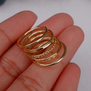 Op de afbeelding: Een verzameling goudkleurige ringen in verschillende ontwerpen. De ringen zijn op elkaar gestapeld en tonen verschillende texturen en stijlen, waaronder gladde banden en bewerkte oppervlakken. De ringen worden gepresenteerd tegen een neutrale achtergrond.