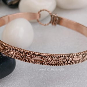 May include: A copper-colored bangle bracelet with an intricate floral pattern. The bracelet features a repeating design of flowers, leaves, and other decorative elements. The bracelet is approximately 0.25 inches wide and has a simple clasp.