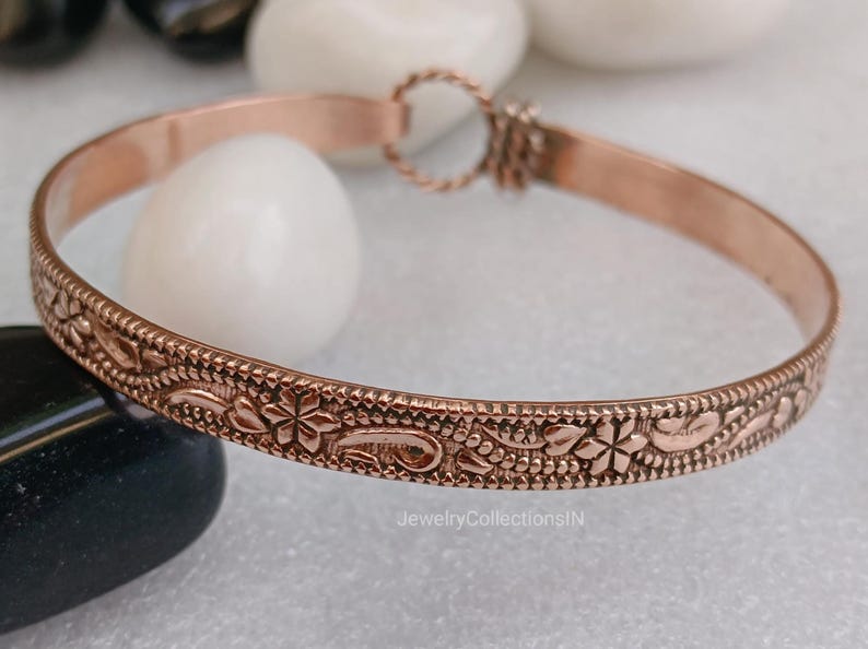 May include: A copper-colored bangle bracelet with an intricate floral pattern. The bracelet features a repeating design of flowers, leaves, and other decorative elements. The bracelet is approximately 0.25 inches wide and has a simple clasp.