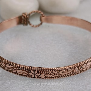 May include: A copper-colored bangle bracelet with an intricate floral pattern. The bracelet features a detailed design of flowers and leaves around the band. The clasp is a twisted rope design. The jewelry is displayed on a light-colored surface.