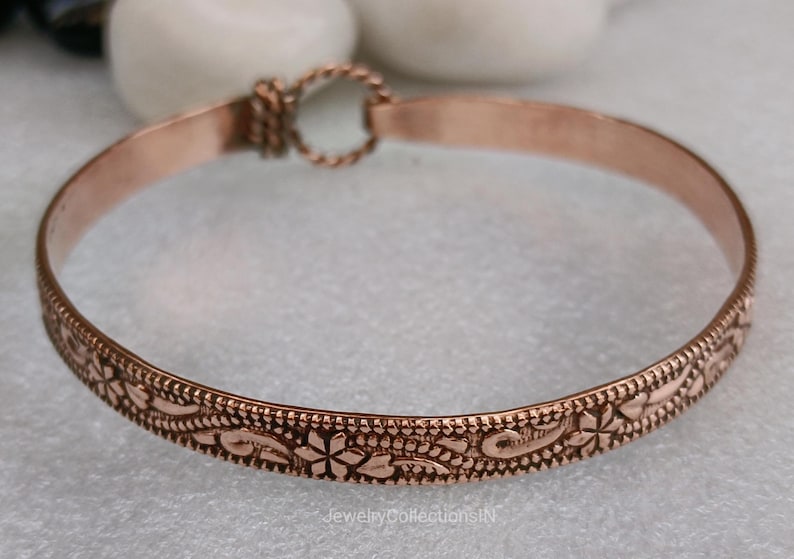 May include: A copper-colored bangle bracelet with an intricate floral pattern. The bracelet features a detailed design of flowers and leaves around the band. The clasp is a twisted rope design. The jewelry is displayed on a light-colored surface.