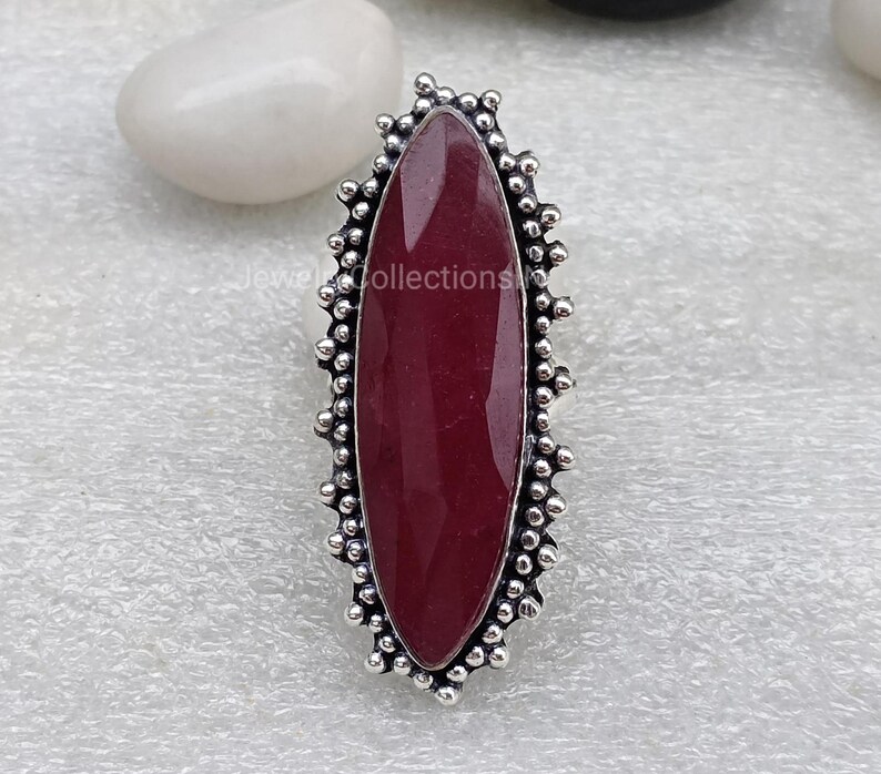 May include: A silver ring featuring a dark red oval gemstone set within a decorative border of small silver beads.