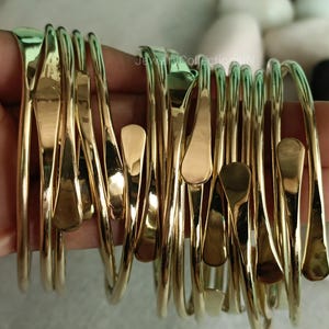 Op de afbeelding: Een set van 12 gouden metalen armbanden met een uniek traanvormig ontwerp. De armbanden zijn gestapeld en in een hand gelegd, waardoor hun ingewikkelde details worden getoond.