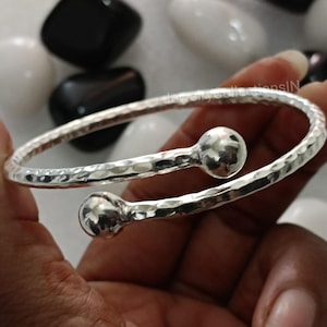 May include: A silver hammered bangle bracelet with two round balls on the ends. The bracelet is open and can be adjusted to fit different wrist sizes.