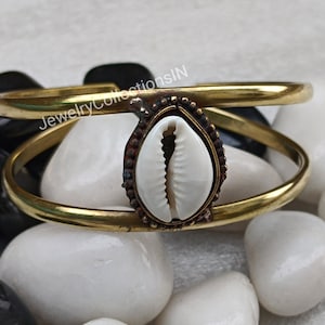 May include: A gold-toned double bangle bracelet with a white cowrie shell charm. The bracelet is made of brass and has a simple, elegant design.