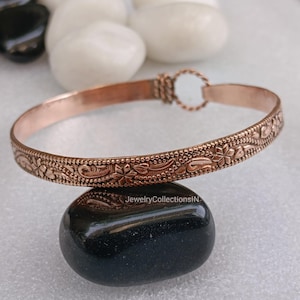 May include: A copper-colored bracelet with a floral pattern. The bracelet is approximately 0.5 inches wide and features a detailed design of flowers and leaves. The clasp is a simple loop and hook closure. The bracelet is displayed on a black stone.