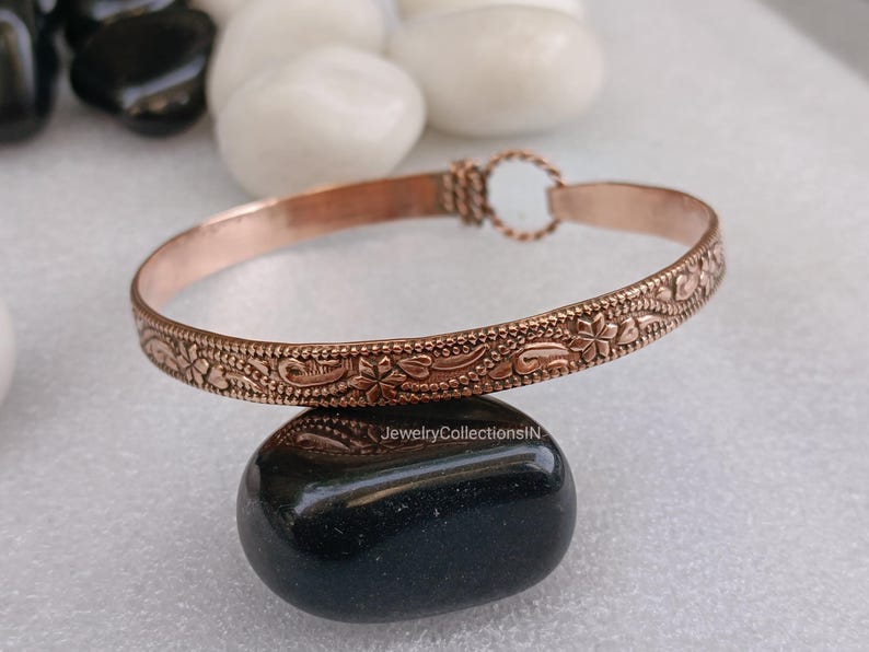 May include: A copper-colored bracelet with a floral pattern. The bracelet is approximately 0.5 inches wide and features a detailed design of flowers and leaves. The clasp is a simple loop and hook closure. The bracelet is displayed on a black stone.