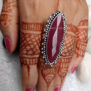 May include: A silver ring featuring a large, oval-shaped, dark red gemstone. The ring has a decorative, beaded design around the edge of the setting.