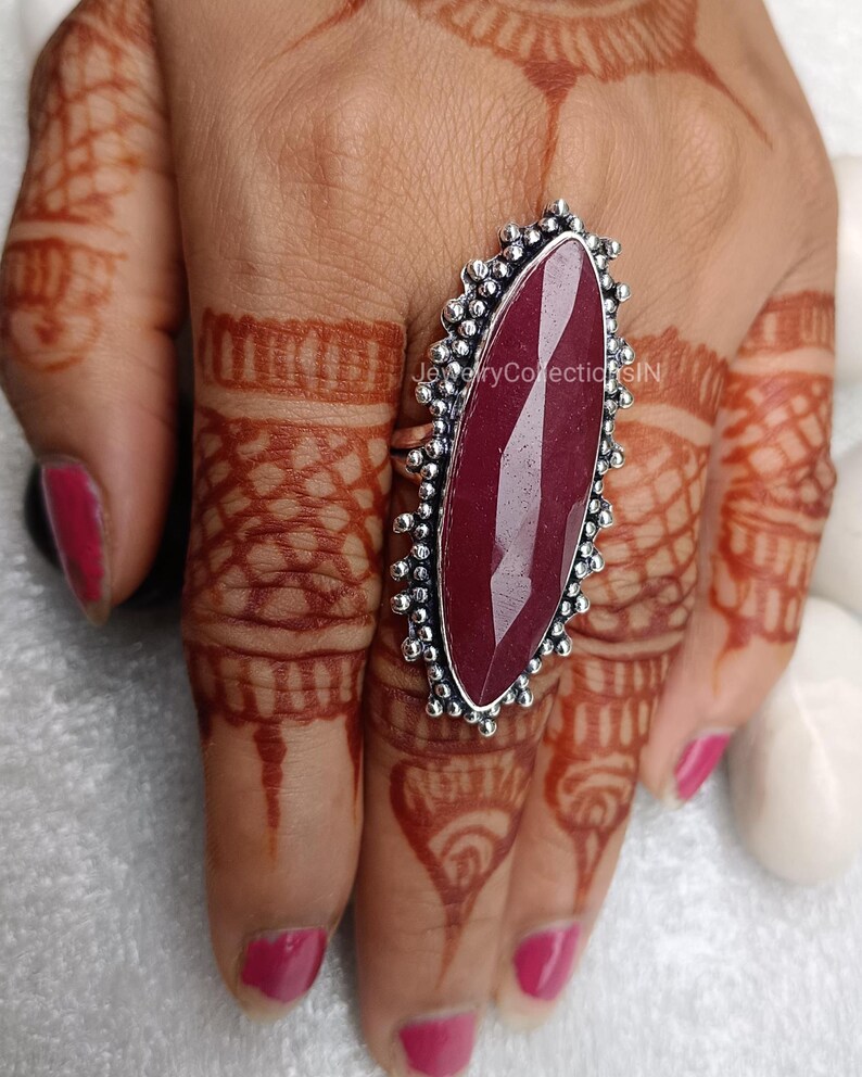 May include: A silver ring featuring a large, oval-shaped, dark red gemstone. The ring has a decorative, beaded design around the edge of the setting.