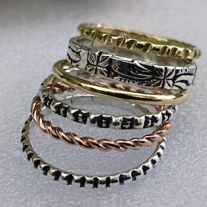 Könnte beinhalten: Ein Satz von fünf Silber- und Goldringen, die übereinander gestapelt sind. Die Ringe haben unterschiedliche Texturen und Muster, darunter ein gedrehtes Seil-Design, ein Ring mit einem Blumenmuster und ein Ring mit einer strukturierten Oberfläche.