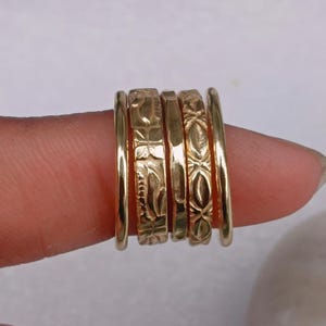 Puede incluir: Un conjunto de anillos dorados, algunos con diseños grabados, apilados en un dedo. Los anillos presentan diferentes patrones, incluyendo estilos geométricos y florales. Se muestran sobre un fondo suave y neutro.