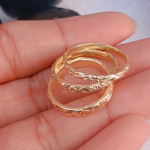 以下が含まれることがあります： 精巧な花柄と幾何学模様が施された、3つの金色の指輪。指輪は重ねられており、その詳細なデザインが際立っています。指輪はおそらく金または金色の金属で作られています。