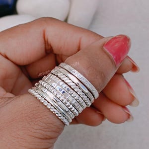 May include: A stack of ten silver rings with different textures and patterns. The rings are worn on a person's thumb.