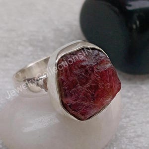 May include: A silver ring with a large, rough-cut, red gemstone. The gemstone is set in a silver bezel setting. The ring is on a white surface.