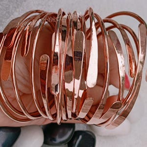 Peut inclure: Un ensemble de 12 bracelets en cuivre, chacun avec une texture martelée et une forme légèrement différente. Les bracelets sont empilés les uns sur les autres, créant un look unique et accrocheur.