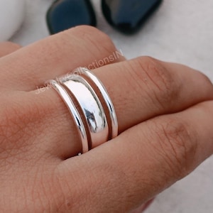 Może przedstawiać: Srebrny pierścionek z trzema pasami ułożonymi jeden na drugim. Paski są gładkie i błyszczące.