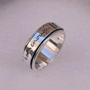 May include: A silver-toned spinner ring with the inscription "I am enough" in a handwritten font. The ring has a textured band and a rotating outer ring. The ring is designed to be worn on a finger.