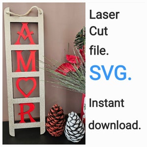 Peut inclure: Une pancarte en bois avec le mot "AMOR" écrit en lettres rouges sur fond blanc. La pancarte est attachée à une corde et a un aspect rustique. La pancarte est devant une décoration de Noël rouge et verte.