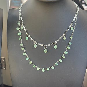 May include: A two-strand necklace with a silver chain and small, teardrop-shaped green gemstones. The necklace is displayed on a black mannequin. The gemstones are evenly spaced along the chain, creating a delicate and elegant design.