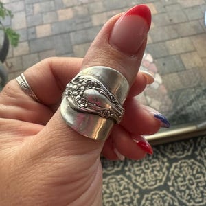 May include: A silver-toned ring crafted from a repurposed spoon, featuring an embossed floral pattern on the front surface. The metal band wraps around the finger, displaying a polished finish with intricate detailing along the central section.